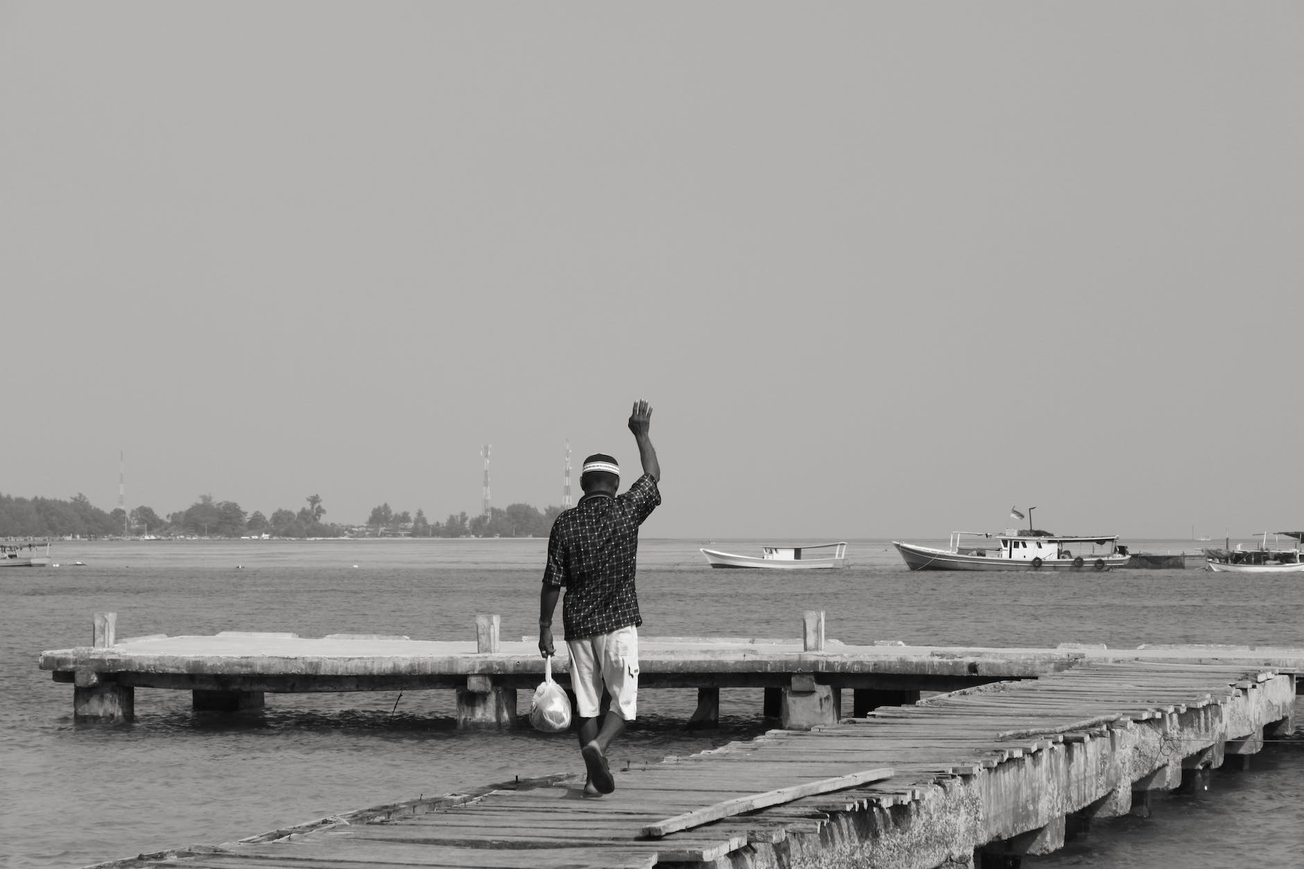 grayscale photo of a man walking on wooden dock waving